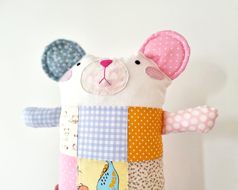 May include: A handmade stuffed bear with a patchwork body. The bear has a white face with pink accents, a pink nose, and two ears, one blue with white polka dots and the other pink with white polka dots. The bear's body is made of various patterned fabrics.