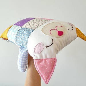 May include: A handmade cat-shaped pillow with a patchwork design. The pillow combines various patterned fabrics in blue, purple, and floral prints. The cat's face is white with pink accents, including a dotted ear and a heart-shaped nose. A charming decorative item.