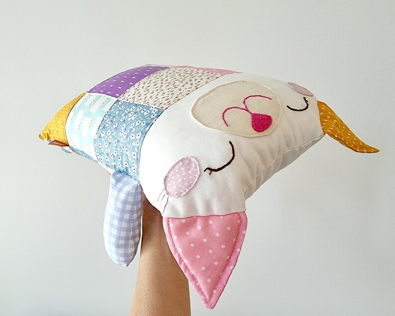 May include: A handmade cat-shaped pillow with a patchwork design. The pillow combines various patterned fabrics in blue, purple, and floral prints. The cat's face is white with pink accents, including a dotted ear and a heart-shaped nose. A charming decorative item.