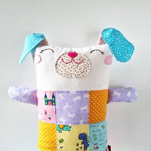 May include: A handmade dog toy with a white face, blue ears, and a patchwork body. The dog's face has a floral nose and stitched eyes. The body is made of various patterned fabrics, including polka dots, rabbits, and castles.