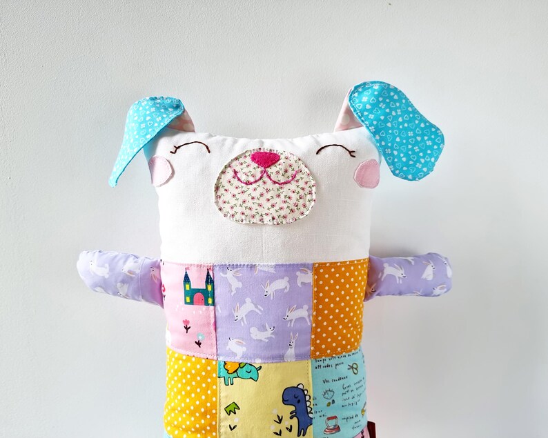 May include: A handmade dog toy with a white face, blue ears, and a patchwork body. The dog's face has a floral nose and stitched eyes. The body is made of various patterned fabrics, including polka dots, rabbits, and castles.