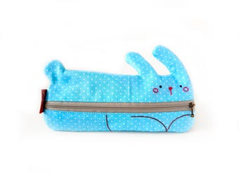 May include: A light blue pencil case with a white polka dot pattern shaped like a rabbit. The case features a horizontal beige zipper closure, embroidered facial details including black eyes and a red nose, and a small red fabric tag on the side.
