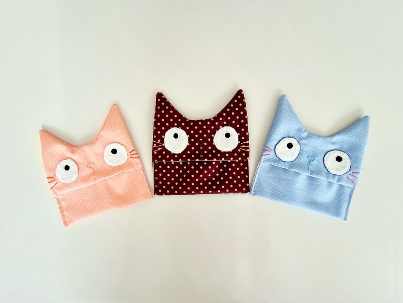 May include: Three fabric cat-shaped pouches with embroidered eyes and whiskers. The pouches are pink with white polka dots, brown with white polka dots, and light blue with white polka dots.