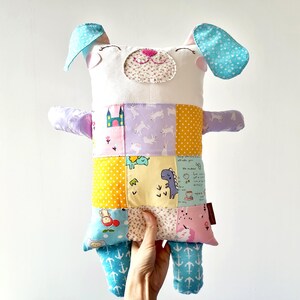 May include: A handmade stuffed dog toy with a patchwork design. The dog has a white head with blue ears and a pink nose. The body is made of colourful fabric squares with various patterns, including castles, dinosaurs, and unicorns.