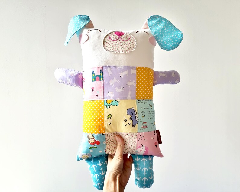 May include: A handmade stuffed dog toy with a patchwork design. The dog has a white head with blue ears and a pink nose. The body is made of colourful fabric squares with various patterns, including castles, dinosaurs, and unicorns.