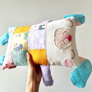 May include: A handmade dog-shaped pillow. The pillow has a patchwork design with various patterns and colours, including yellow polka dots, blue whales, and purple rabbits. The dog's face has floral eye details and pink accents.
