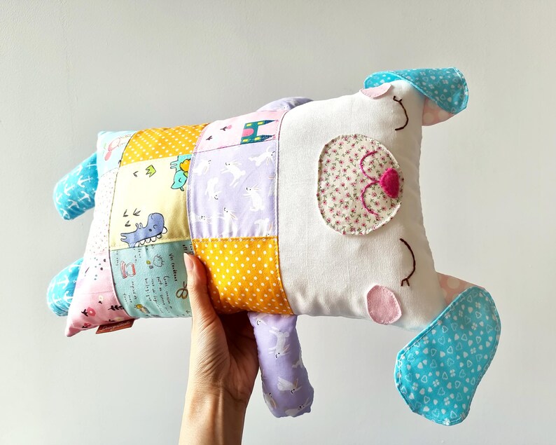 May include: A handmade dog-shaped pillow. The pillow has a patchwork design with various patterns and colours, including yellow polka dots, blue whales, and purple rabbits. The dog's face has floral eye details and pink accents.