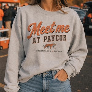 May include: A light gray crewneck sweatshirt featuring the orange and black text Meet me AT PAYCOR, a graphic of a tiger, and the text CINCINNATI, OHIO EST. 1968. The sweatshirt has long sleeves with ribbed cuffs and a ribbed neckline.