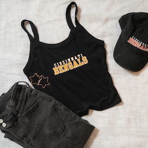 May include: A black tank top with the text CINCINNATI BENGALS in orange and white, a black baseball cap with the same text, a pair of dark gray jeans, and two gold-toned star-shaped earrings arranged on a light-colored fabric surface.
