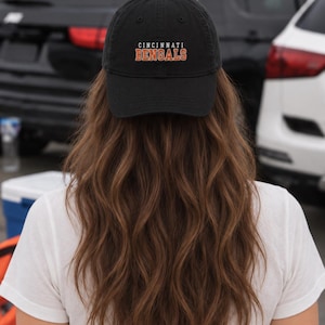Könnte beinhalten: Eine schwarze Baseballkappe mit dem Schriftzug CINCINNATI BENGALS, der in weißen und orangefarbenen Blockbuchstaben auf der Vorderseite eingestickt ist. Die Kappe hat eine strukturierte Krone, einen gebogenen Schirm und einen Knopf oben.