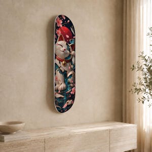 May include: A skateboard deck featuring a graphic of a white beckoning cat with blue and gold kintsugi-style cracks, a red collar with a gold bell, pink cherry blossoms, and a large red sun against a dark background.