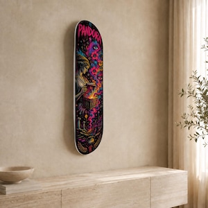 Op de afbeelding: Een skateboarddeck met een kleurrijke afbeelding van een figuur die een doos opent met het opschrift PANDORA. Het kunstwerk bevat wervelende roze, blauwe en oranje rook, gestileerde bloemen en ogen tegen een zwarte achtergrond.
