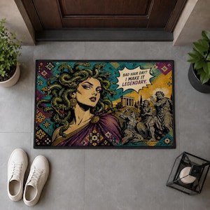 Op de afbeelding: Een rechthoekige deurmat met een stripachtige illustratie van Medusa met groene slangen als haar, gekleed in een paars gewaad. Een tekstballon bevat de tekst: BAD HAIR DAY? I MAKE IT LEGENDARY. Op de achtergrond zijn antieke ruïnes te zien.