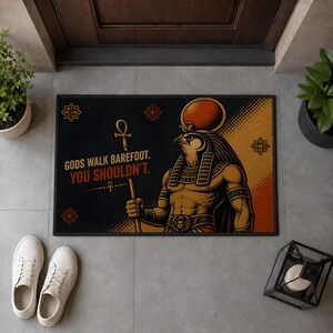 Puede incluir: Un felpudo rectangular con fondo negro y una ilustración gráfica de la deidad egipcia Ra en tonos dorados y naranjas. El texto dice "GODS WALK BAREFOOT. YOU SHOULDN'T." junto a un símbolo ankh y motivos florales decorativos.