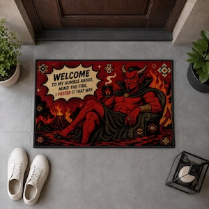 May include: A rectangular doormat featuring a red devil sitting in a chair, holding a mug, with a volcano and flames in the background. A speech bubble contains the text: WELCOME TO MY HUMBLE ABODE. MIND THE FIRE, I PREFER IT THAT WAY.
