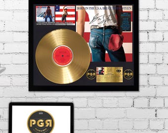 May include: A gold record award plaque with a black frame. The plaque features a gold record, a photo of Bruce Springsteen, and the album title "Born in the U.S.A." The plaque also includes a signature from Bruce Springsteen and a platinum and gold record company logo.