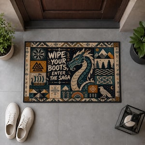 Könnte beinhalten: Eine rechteckige Fußmatte mit einer blauen Drachenillustration und dem Text "WIPE YOUR BOOTS, ENTER THE SAGA". Das Design zeigt nordisch inspirierte Motive wie ein Wikingerschiff, einen Raben, Berge und geometrische Muster in Blau, Gold und Creme.
