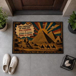 May include: A rectangular doormat featuring a graphic of the Great Sphinx and pyramids of Giza with camels. The text reads: BUILT THOUSANDS OF YEARS AGO, STILL STANDING. WITNESS TO HISTORY. ETERNAL AS LEGENDS. The design uses a vintage sunburst background.