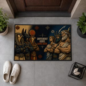 May include: A rectangular doormat featuring a graphic illustration of Egyptian deities on the left and Greek gods on the right. The text in the centre reads DIFFERENT GODS. SAME DOOR. The design uses a colour palette of gold, blue, and black.