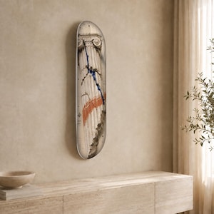 May include: A skateboard deck featuring a graphic print of a weathered classical stone column with a Corinthian capital. The design includes a prominent vertical blue streak and a horizontal orange brushstroke across the textured stone surface.