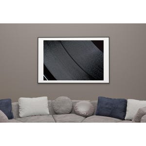 Op de afbeelding: Een ingelijste abstracte kunstafdruk met een close-up van een getextureerd, zwart oppervlak met een gebogen lijn. Het kunstwerk wordt tentoongesteld op een grijze muur boven een grijze bank met diverse kussens.