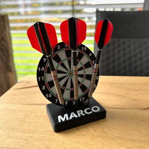Könnte beinhalten: Eine Dartscheibe mit drei Darts, mit roten und schwarzen Flights, wird auf einem Holztisch präsentiert. Die Dartscheibe ist schwarz und weiß mit Zahlen. Der Sockel ist schwarz mit dem Namen "MARCO" in Weiß.