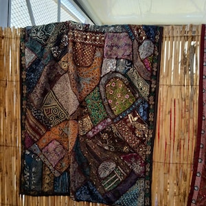 May include: A colourful patchwork tapestry with intricate embroidery and sequins. The tapestry is made of various fabrics in shades of brown, purple, green, and blue. It is hanging on a bamboo wall.