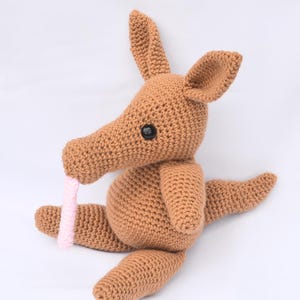 Può includere: Un peluche di oritteropo all'uncinetto marrone, con un lungo muso, grandi orecchie e una lingua rosa. L'oritteropo ha occhi neri a bottone ed è seduto con le braccia e le gambe distese.