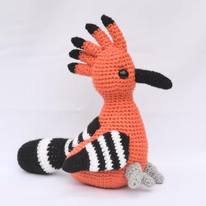 May include: A handmade crochet bird in shades of orange, black, white, and grey. The bird has a distinctive crest, a long beak, and striped wings and tail. The bird is sitting on a white surface.