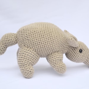 Può includere: Un giocattolo di peluche di formichiere all'uncinetto di colore beige chiaro. L'animale di peluche fatto a mano ha un lungo muso, piccole orecchie e occhi neri. Il formichiere è posizionato a quattro zampe, con la coda estesa.
