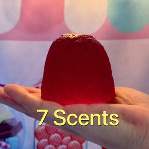 May include: A large, translucent, red, jelly-like object is held in a hand. The text "7 Scents" is displayed in yellow, indicating a product with multiple fragrances. The background features a pink and white pattern.