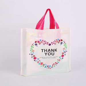 May include: A white plastic shopping bag with pink handles. The bag features a heart-shaped floral design with the words "THANK YOU" in the center. The bag also has a quote at the bottom.