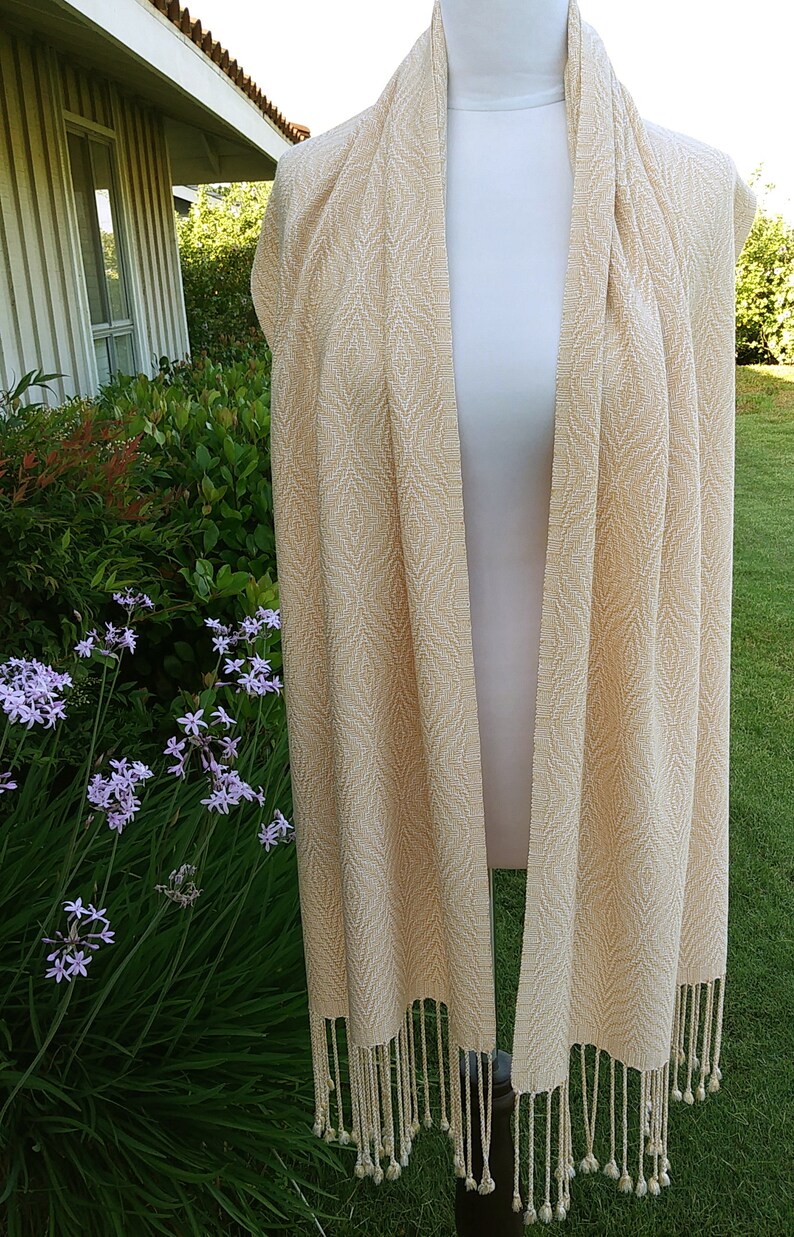May include: A cream-colored woven scarf with a subtle geometric pattern and fringe tassels.