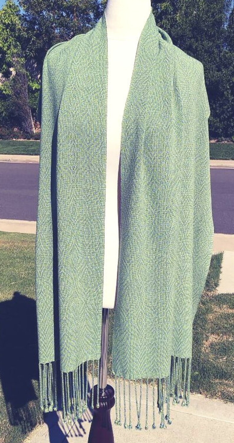 May include: A green and white woven shawl with a fringe border. The shawl is draped over a mannequin.
