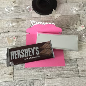 May include: Pink card with a white cutout of the word "Mom" decorated with rhinestones. A silver envelope and a Hershey's milk chocolate bar are placed on top of the card.