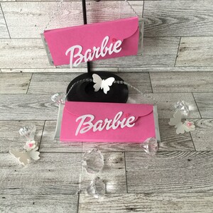 May include: Two pink cardboard boxes with the word "Barbie" printed in white on them. The boxes are decorated with white butterflies and pink rhinestones.