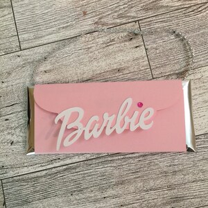May include: A pink cardboard box with a silver border and a white "Barbie" logo. The box has a flap on the top and a pink gem on the front.