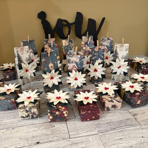 May include: A collection of rectangular gift boxes wrapped in various dark-toned holiday-themed paper, each topped with a cream-colored paper poinsettia featuring three small red beads in the center and secured with a wooden clothespin.