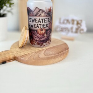 May include: A frosted glass can-shaped tumbler with a wooden lid, featuring a fall-themed design. The design includes pumpkins, knitted sweaters, and the words "Sweater Weather". The tumbler sits on a wooden cutting board.