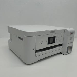 May include: A white Epson printer with a flat top and a control panel featuring a small screen and several buttons. The printer has a paper tray and ink cartridge access. The Epson logo is visible on the top.