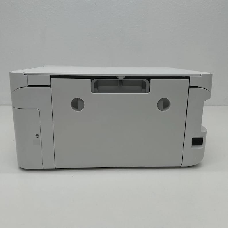 May include: A white printer with a flat top and a grey handle. The printer has a rectangular shape with rounded corners and a small power button on the side. The printer is designed for home or office use.