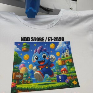 May include: White t-shirt with a vibrant graphic print of a blue cartoon character in a video game scene. The design includes coins, a castle, and the text "NBD STORE / ET-2850".