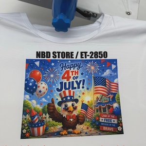 May include: White t-shirt with a colourful graphic print for the 4th of July. The design features an eagle wearing a patriotic hat, fireworks, American flags, and the text "Happy 4th of July!" The text "NBD STORE / ET-2850" is also visible.