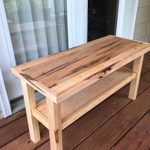 May include: A light-colored wooden bench with a lower shelf. The bench has a rectangular top with a natural wood grain pattern and dark streaks. The legs and shelf are made of lighter wood. The bench is approximately 48 inches long, 16 inches wide, and 18 inches high.