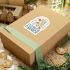 May include: A brown gift box with a light green ribbon and a sticker that reads "Flower Farmer" with orange flowers. The sticker also says "Support Your Local". The box is surrounded by Christmas decorations.