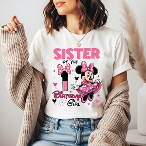 May include: White t-shirt with a pink graphic that says "SISTER OF THE BIRTHDAY GIRL". The design includes a number 1, Minnie Mouse, bows, and stars. The shirt is a casual style.