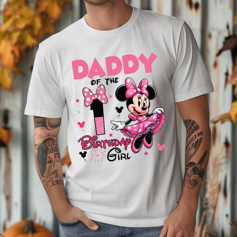 May include: White t-shirt with a pink graphic featuring the text "DADDY of the BIRTHDAY GIRL" and a Minnie Mouse design. The design includes a large number 1 and a bow. The shirt is a crew neck.