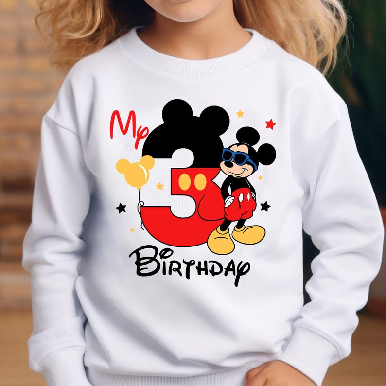 May include: White sweatshirt featuring a Mickey Mouse design for a third birthday. The design includes a large red number 3, Mickey Mouse wearing sunglasses, and the words "My Birthday". Yellow and red accents complete the design.