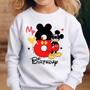 May include: White sweatshirt featuring a colorful Mickey Mouse design. The design includes a large number 8, the words "My Birthday", and a cartoon Mickey Mouse character wearing sunglasses. The design also includes stars and a balloon.