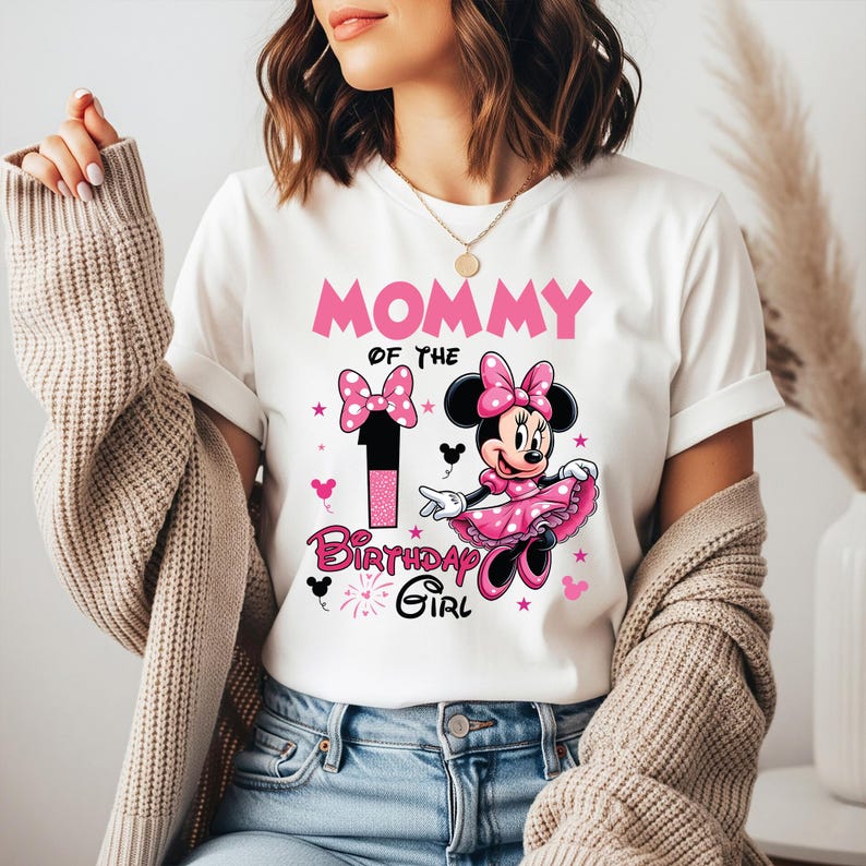 May include: White t-shirt with the text "MOMMY OF THE BIRTHDAY GIRL" in pink. The shirt features a graphic of Minnie Mouse in a pink dress and bow, along with a number 1 and pink stars. A fun and festive design.
